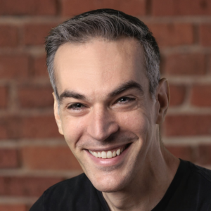Dan Shapiro Crop Headshot of a smiling person with short dark hair, wearing a dark shirt in front of a brick wall background.