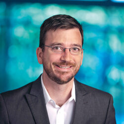 Headshot of a person wearing glasses and a suit jacket with a white shirt, smiling in front of a blurred blue-green background.