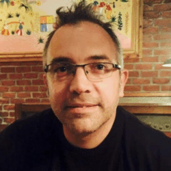 Headshot of a person wearing glasses, in front of a colorful picture and brick wall.